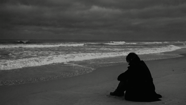 A solitary figure sits on the beach, gazing at the turbulent waves under a cloudy sky.
