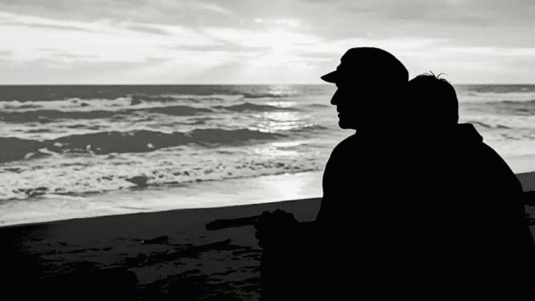 Silhouettes of two people sitting close together on a beach, gazing at the ocean under a cloudy sky.
