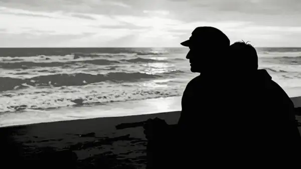 Silhouettes of two people sitting close together on a beach, gazing at the ocean under a cloudy sky.